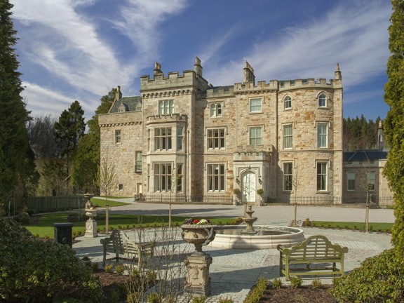 An exterior view of Crossbasket Castle shows a large 3 storey blonde sandstone building with large bay windows, crenelated walls and carved details. A paved pathway leads to the building with a fountain, benches and stone ornaments in the foreground. Lawns, flower beds and trees surround the building.