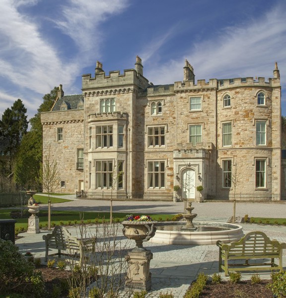 An exterior view of Crossbasket Castle shows a large 3 storey blonde sandstone building with large bay windows, crenelated walls and carved details. A paved pathway leads to the building with a fountain, benches and stone ornaments in the foreground. Lawns, flower beds and trees surround the building.