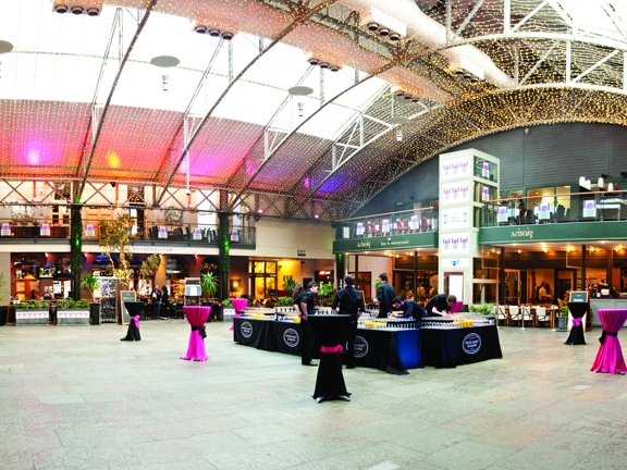 An interior view of Merchant Square, a large space with a paved stone floor and a curved roof, with skylights. The photo is taken from the ground floor during the day. Furniture is arranged for an event, four tables in a square are covered in glasses of juice or wine; tall tables dot the open space. Around the edge, fenced off seating areas and the entrances to several businesses are visible. There is a  glass lift between the ground floor and the first floor balcony in the centre of the far wall.