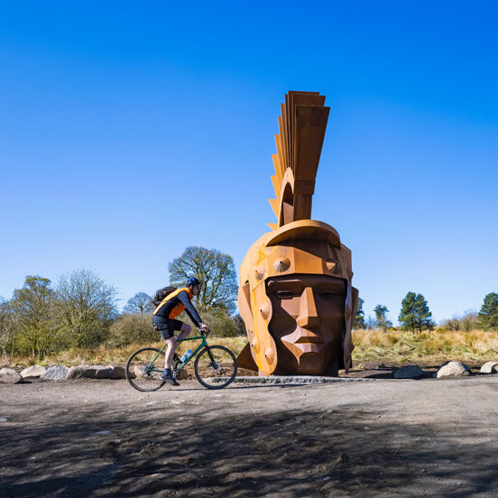 Cyclist passing in front of the 6m tall metal sculpture of the head of roman soldier