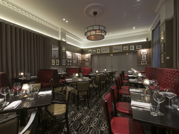 An Anchor Line private dining room with a dark, ring-patterned carpet. The ceiling is white with plaster work beading and a central ceiling rose around the modern cylindrical light fitting. The room is filled with small dark dining tables; with chairs, benches and booths in red and taupe leather arranged around them. The far wall has a wide, wooden, concertina door, most of its width, it is closed off to separate the room. A wide, heavy, brown curtain covers much of the left wall suggests a window.