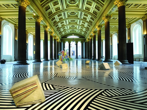 An interior shot of The Gallery of Modern Arts, Gallery 1. The image shows a large room lined with large arched windows. 2 rows of dark Roman-style columns line the room, reaching up to the gold, arched ceiling covered in decorative plaster work. The art installation covers the floor in a striped black and white pattern and concrete shapes and a colourful, wiry sculpture can be seen in the distance. Spotlights can be seen between the columns and the outer walls are uplit.