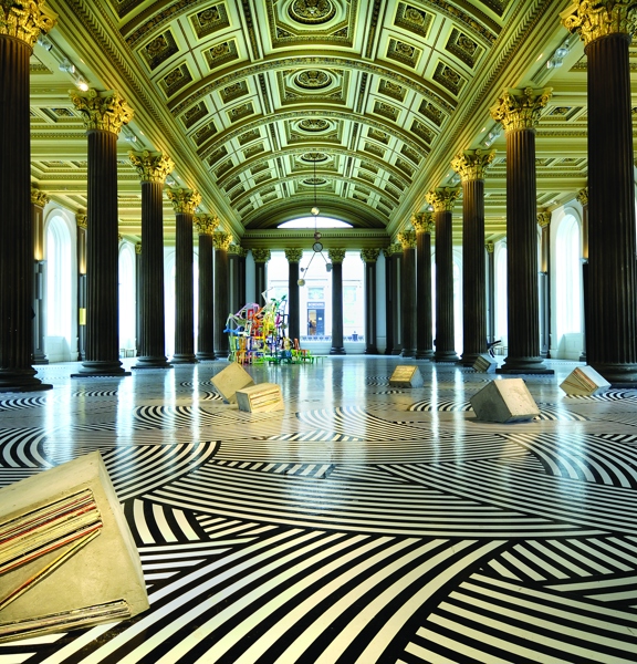 An interior shot of The Gallery of Modern Arts, Gallery 1. The image shows a large room lined with large arched windows. 2 rows of dark Roman-style columns line the room, reaching up to the gold, arched ceiling covered in decorative plaster work. The art installation covers the floor in a striped black and white pattern and concrete shapes and a colourful, wiry sculpture can be seen in the distance. Spotlights can be seen between the columns and the outer walls are uplit.