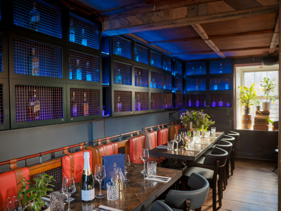An interior shot of The Finnieston restaurant dining room. A narrow room with floorboards and dark walls, the left hand wall is lined with cubby holes with metal grills over them, they're lit blue and hold bottles of spirits. A long wooden bench runs the length of this wall, there are red leather cushions on the backs and seats. 2 rectangular, tiled dining tables are visible, they are laid and decorated with menus, potted plants, wine bottles & more. The window in the far wall has potted plans on its ledge.