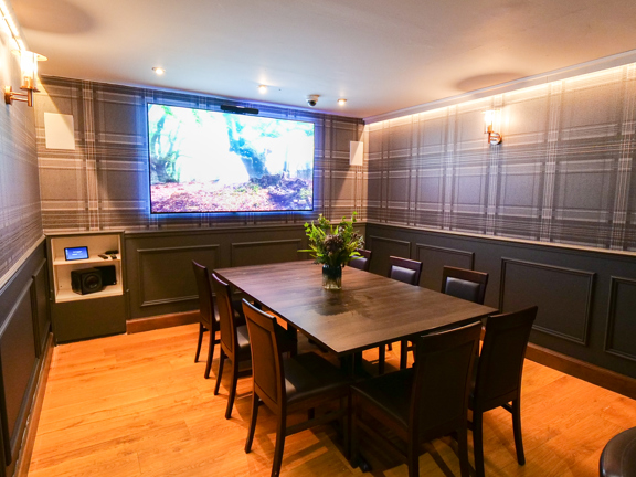 An interior of a small private dining space at Mharsanta, shows a room decorated with wooden floorboards, dark panelling and grey, tartan wallpaper. Dark wood and leather chairs can be seen around a dark, rectangular dining table, with a vase of flowers in its centre. The far wall is dominated by a large television screen which lights the room. Small wall sconces and spotlights also do. A small shelf in the bottom left corner of the wall seems to house a tablet, remotes and a speaker.