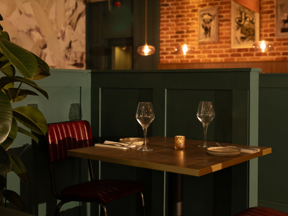 A close quarter interior detail of a table laid for dinner at Table Twenty Eight. The wooden table is positioned against a panelled half-wall, painted a chic grey-green. Seats, facing each other across the table are chrome with red leather padded seats and backs. A large, dark green rubber plant sits to the right of the table. Beyond the half wall, statement wallpaper and bare brick walls bare black and white prints. Modern glass lightshade hang from the ceiling.