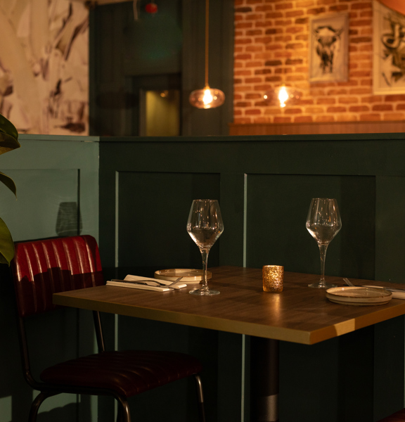 A close quarter interior detail of a table laid for dinner at Table Twenty Eight. The wooden table is positioned against a panelled half-wall, painted a chic grey-green. Seats, facing each other across the table are chrome with red leather padded seats and backs. A large, dark green rubber plant sits to the right of the table. Beyond the half wall, statement wallpaper and bare brick walls bare black and white prints. Modern glass lightshade hang from the ceiling.