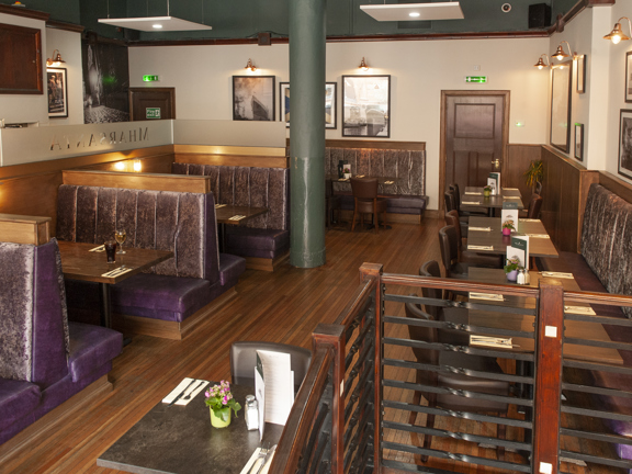 An interior view of Mharsanta shows a restaurant with dark-wood floors and white walls, decorated with framed photos. Square, polished-stone or wooden topped tables are arranged in booths and the surrounding space. The booths and benches that line the room are upholstered in purple leather and velvet and the chairs are dark wood and leather. Dark doors with fire exit signs can be seen in either corner of the image. A banister in the foreground suggests there are stairs down from the main restaurant floor,  