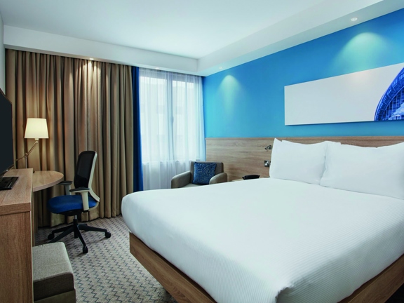 An interior view of a Hampton by Hilton bedroom, the room is decorated with a zigzag patterned carpet in neutral tones and the wall behind the bed is a bright turquoise-blue; the remaining walls are white and the furnishing are wooden. The bed on the right is made with crisp, white sheets. A television is on a surface at the foot of the bed, a desk and chair are in the far left corner and an armchair sits in the window in the back right corner. An abstract print of the SEC armadillo is over the bed.