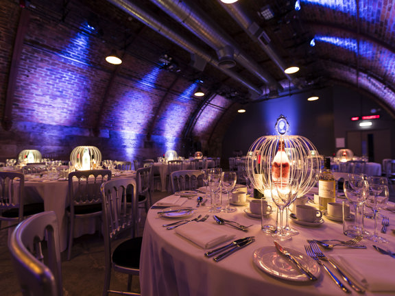 An interior image from Platform set up for an event, shows an arched, cavernous brick tunnel, the floor that is visible between the tables looks like concrete. The space is filled with round tables with white table cloths. Silverware, wine glasses and glowing, lantern centre pieces decorate them. They are surrounded by silver chairs with velvet seats. The space is dimly lit, the walls are lit with blue spotlights and white pendant lights hang from the curved, brick ceiling.
