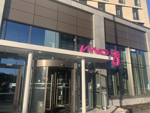 An exterior shot of Moxy SEC's main entrance shows a glass revolving door, with an automatic, assisted-door on either side, the flooring is a mix of tarmac and paved.  A white metallic awning above supports 3D, bright pink, wiggly lettering that reads, "moxy". The ground floor is windowed and the floors above are rendered in cream with dark-framed modern windows. A sliver of blue sky is visible in the top-right corner and blue sky and a tall building are reflected in the ground-floor windows.