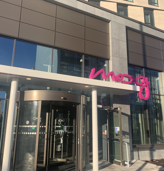 An exterior shot of Moxy SEC's main entrance shows a glass revolving door, with an automatic, assisted-door on either side, the flooring is a mix of tarmac and paved.  A white metallic awning above supports 3D, bright pink, wiggly lettering that reads, "moxy". The ground floor is windowed and the floors above are rendered in cream with dark-framed modern windows. A sliver of blue sky is visible in the top-right corner and blue sky and a tall building are reflected in the ground-floor windows.