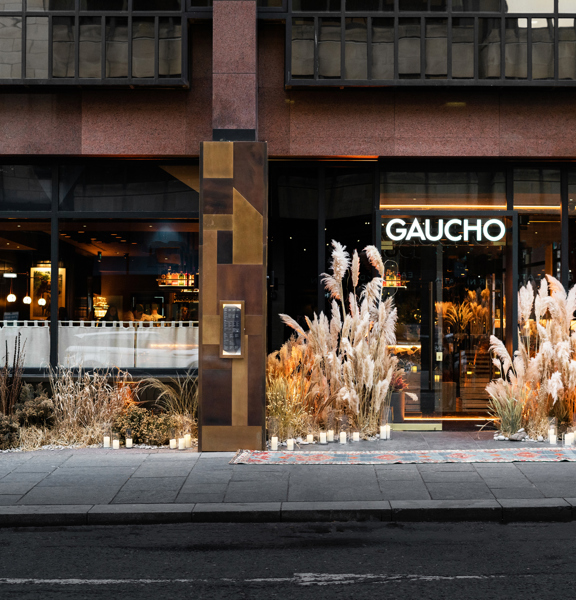An exterior shot of the restaurant Gaucho shows the ground floor entrance of a modern building in shades of polished red stone, with plate glass windows and glass doors. White, lit letters read, "GAUCHO" above the door. Metallic panels of brass and copper frame the doorways, with small inset windows housing menus. The entrance is further decorated with a sweeping landscape of dried grasses, foliage, plants and candles. The pavement is wide and paved with slabs, there is a small curb into the tarmacked road.