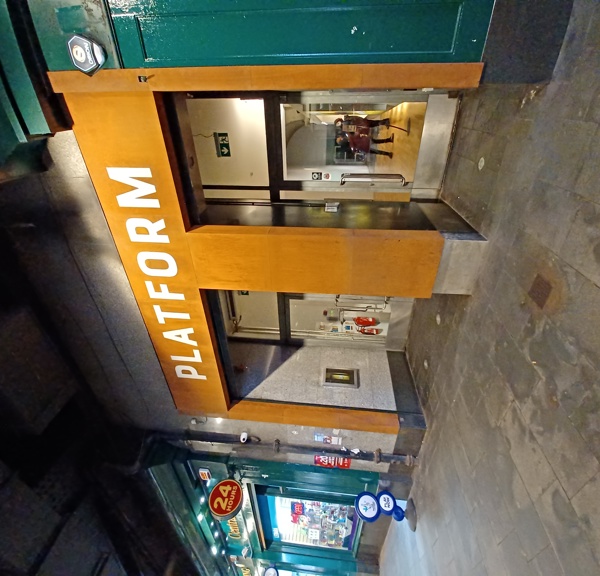 The front entrance to Platform Glasgow shows a blocky, concrete entranceway with glass doors and polished wooden facing. The entrance is sandwiched between two traditional, store fronts painted with green glossy paint. The pavement outside is wide and paved and has water marks. White lettering on the varnished wood facing reads "PLATFORM". It is possible to see wooden floors inside.