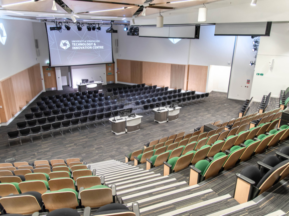 A photo from the taken from the height of the tiered seating inside a large, bright, modern lecture theatre at the TIC. The outer walls are angled coming to meet at the projector screen that dominates the front of the room. The tiered seats are green and self-righting, further grey chairs are arranged in rows on the flat floor at the front of the room. The floor and stairs are carpeted. Technical equipment including lighting rigs, speakers, projectors and lecterns with mics & monitors are notable.
