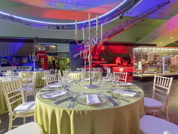 The Riverside Museums main hall arranged for a private dinner. Glass display cases, vehicles and a telephone box are seen around the periphery of the space & lit with coloured spotlights. Round tables are decorated with yellow cloths, tall silver candelabras, glasses and silverware. Cream, spindle back chairs with padded seats surround them. Where the floor is visible it looks like polished concrete.