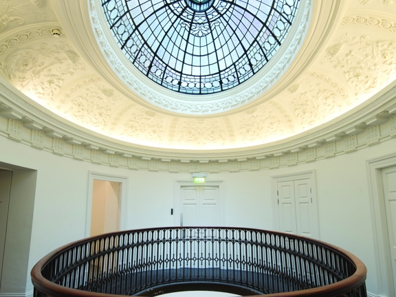 An interior view from GoMA shows an oval space with a balcony in the centre of it. The landing is carpeted and has white walls. The balcony has an ornate black railing and a dark wooden bannister running around it. Various doorways can be seen around the perimeter. The ceiling has an ornate leaded glass oval, skylight at its centre, surrounded by a wide band of white floral plaster work.