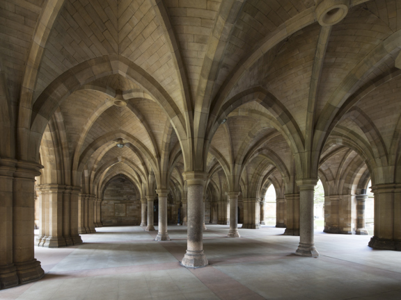 An image of Glasgow University's famous cloisters. Sandstone pillars of different sizes, arches and vaulted ceilings from a mesmerising geometric pattern. The floor is even, stone paving. The photo is taken in the day and daylight can be seen between some of the pillars.