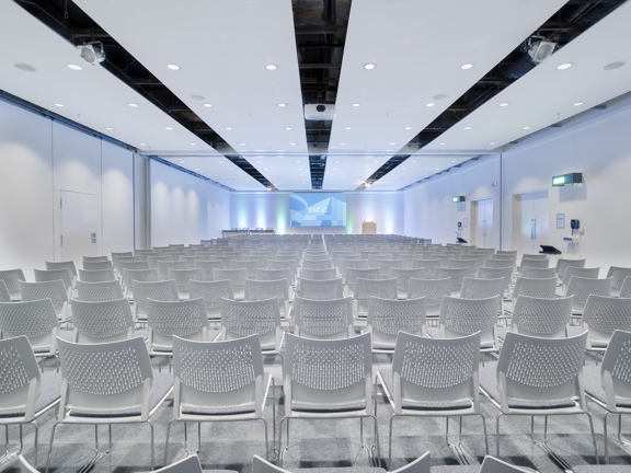 A view of an interior from the SEC shows a large bright white room filled with 20+ rows of white molded plastic, wire legged chairs. The room is carpeted with a very pale grey carpet; the furthest bright, white wall is uplit with icy blue lights. A large projection screen, bearing a blue SEC branded slide is centred; a white lectern and a panel table flank it on either side. Technical lighting and a projector are mounted on the ceiling. 2 fire exits an be seen on the right hand wall.