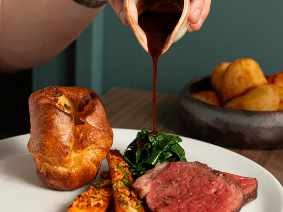 A close up image of a Table Twenty Eight roast dinner. A white, masculine hand and fore-arm can be seen pouring gravy from a small ceramic jug onto a clean plate of roasted vegetables, sliced beef and Yorkshire pudding. A rustic ceramic bowl of roast potatoes can be seen on the wooden table against grey-green panelling. 