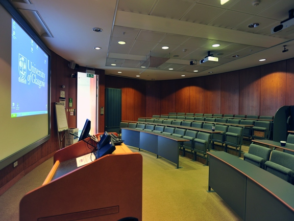 A small lecture theatre at the University of Glasgow. A small, dimly lit space with 4 rows of tiered, self-righting seats. The floor is carpeted and the walls are panelled with wood. The image is taken from the front corner of the space and the edge of a wooden lectern, a monitor, projection screen and flipchart can all be seen. 2 projectors are mounted on the ceiling, further tech equipment is visible at the back of the room. On the fire side of the room an open Fire Exit lets daylight in.