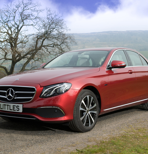 An image of a metallic red Mercedes parked on a gravel road. A verdant, hilly landscape and blue cloudy sky behind is framed by bare trees on either side. The car's license plate reads "LITTLES". 