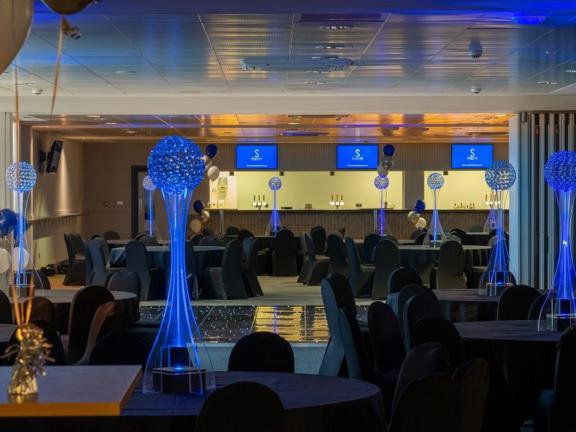 A view of the interior of Hampden Stadium's Bell & Baird event suite. The space is darkened to show the lit dancefloor and perspex centrepieces on the tables. The space is otherwise lit by the light from the bar, which stretches the length of the far wall. There is also light from the 3 television screens on the wall above the bar. The round tables and chairs are covered in black covers and balloon arrangements can be seen throughout the space.