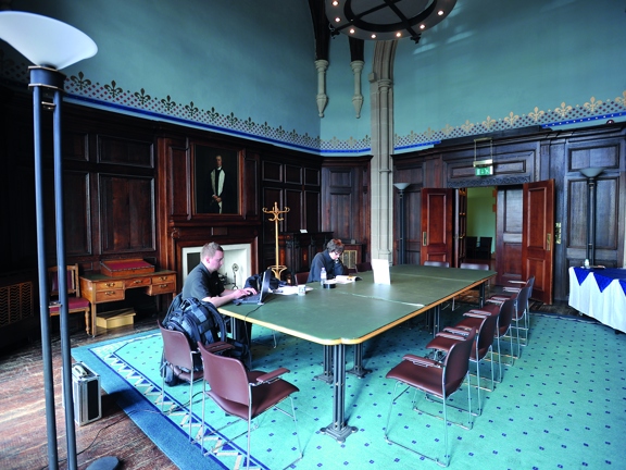 The UoG's Turnbull Room shows a room with dark wooden floor boards, covered in main part, with a thick, patterned, turquoise rug. 3 large tables are placed together, with 14 wire-legged chairs around them, 2 seats are taken by people working. The room is old and lavish with dark wooden panelling and a large carved stone archway in the far wall. The upper portion of the walls are painted a matt turquoise with a border of gilt fleur-de-lis. A white stone fireplace and a large oil portrait adorn the left wall.