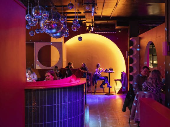 An interior view from August House shows a vibrant bar scene with a mix of high and low seats and tables. A large accent wall at the end of the room has a large circle set into the wall and lit orange. Other arched and round details cut out of walls repeat the motif. Disco balls hang from the ceiling, clustered together, throwing specs of blue light over the otherwise orange lit space. The floor looks smooth and polished.