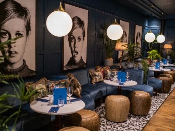 An image of the 63rd & 1st's bar. 2 low navy velvet, C-shaped benches sit side by side, covering the length of the left wall. The walls are also painted dark blue and are panelled. 4 large black & white prints of the supermodel Twiggy punctuate the length. Low, round white tables and leather footstools form small gathering of seats. The floor to the right of the image is wooden, but under the seats and tables is tiled in a patterned, monochrome design. Lamps and varied potted plants decorate the space.