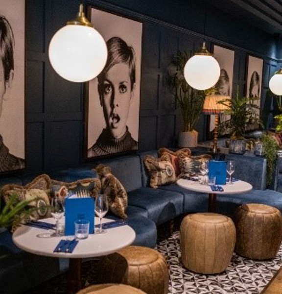 An image of the 63rd & 1st's bar. 2 low navy velvet, C-shaped benches sit side by side, covering the length of the left wall. The walls are also painted dark blue and are panelled. 4 large black & white prints of the supermodel Twiggy punctuate the length. Low, round white tables and leather footstools form small gathering of seats. The floor to the right of the image is wooden, but under the seats and tables is tiled in a patterned, monochrome design. Lamps and varied potted plants decorate the space.