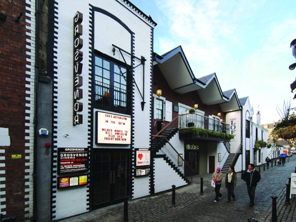 An exterior view of the Grosvenor cafe and cinema shows a white rendered building with black-painted brick details, it boasts features like first-storey windows and pulleys from the buildings original purpose. An entrance on the left hand side has a lit sign reading "Grosvenor," vertically, there is also a lit board with film listings and further posters below. 2 symmetrical staircases lead to a balcony, beside the stairs there is text reading, "The Loft." The building is on a cobbled lane with bollards.