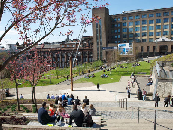 A view across the Strathclyde University's Rottenrow Gardens shows a steep landscaped area with paved walkways, stone steps, lawns and trees. Students can be seen walking across the garden or sitting on stone steps and benches enjoying the sunshine. Modern lampposts and public bins for waste and recycling can be seen on different levels of the space.  A street runs along the far edge of the garden, a parked car and a large brick University building can be seen on it against a blue sky.
