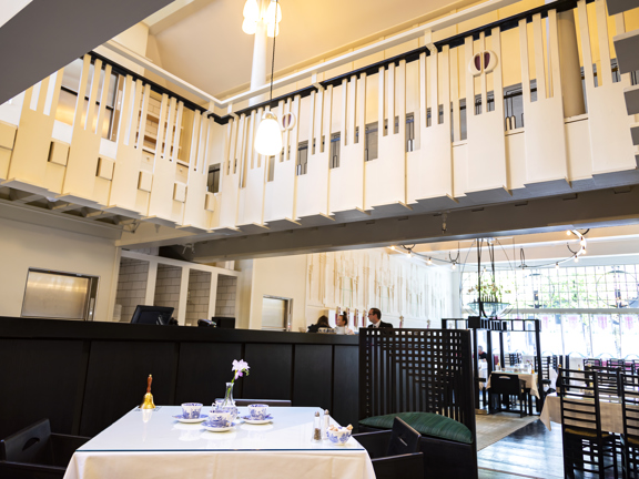An interior view of the Mackintosh at The Willow tearoom, the view is from the ground floor looking up at the first storey balcony. The banister is white and slatted with a repeating, mauve, stylised-floral motif. A square table with 2 low backed black-wood chairs is arranged with blue and white Willow pattern china. To the right of the image is a view of the seating area and window at the front of the venue. The chairs have tall, laddered backs. A Glasgow-Style mural and stained glass can also be made out.