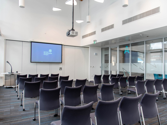 An image of the interior of a small, private meeting space at the TIC. Dark fabric chairs with metal legs are arranged into several rows. They face a projector screen displaying the Strathclyde University logo. The room is decorated with dark striped carpet and white walls. A projector, modern light fittings and vents are fitted in the ceiling. The light in the room suggests the left wall is windowed, the right, interior wall is also glass. It's not possible to see through because of reflections.
