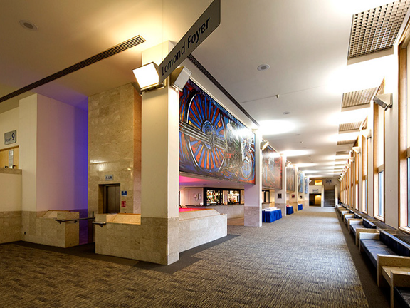 An interior image of the GRCH's Lomond Foyer shows a long, high-ceilinged corridor. A large, colourful fresco decorates the top half of the wall on the left of the corridor, while square pillars with polished stone bases punctuate the length. A long bar can be seen behind the pillars. On the right, exterior wall, windows run the length of the corridor with angular leather benches and polished stone surfaces just in front of them. A lift, a set of stairs and 2 doorways are also visible off of the foyer.