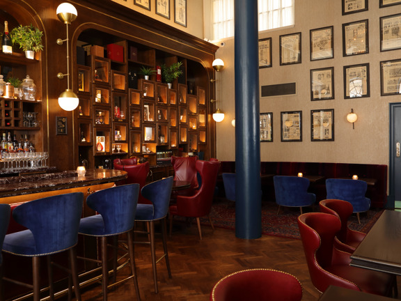 An interior of The Citizen's, Editors Suite bar. The floor is polished, herringbone parquet, (a rug can be seen under some of the tables at the end of the room.) The walls are cream with framed newspaper pages. The left wall, behind a long wooden and veined marble bar is made up of varnished wooden pigeon hole shelves. The shelves are lit and contain, glasses, bottles, plants and more. The seating is a mixture of chairs and high stools, they are leather or velvet in rich shades of royal blue and crimson.