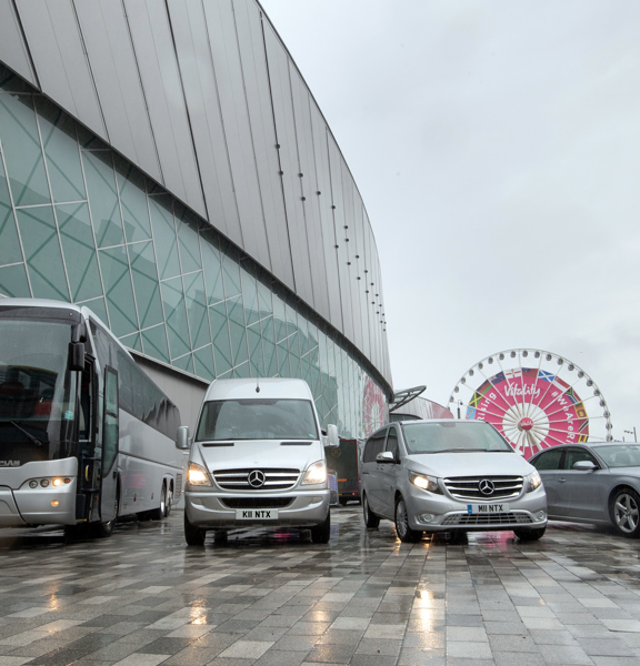 A silver coach, van, people carrier and car are parked in a row besides the silver, curving wall of a large modern building. The ground is paved with stone slabs, wet from rain. The sky is grey and the cars headlights are on. A white ferriswheel with pink branding, blue boarding and 2 modern, brick buildings can all be see in the background.