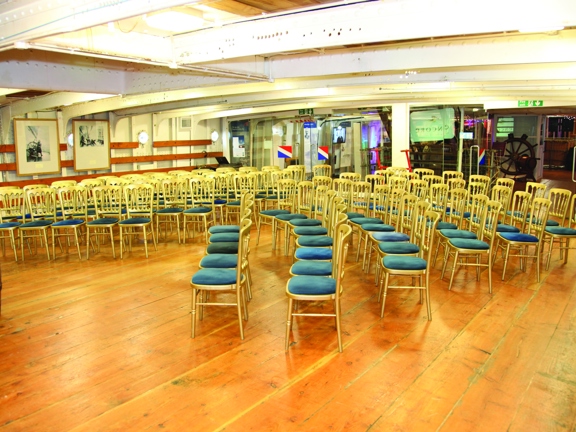An interior from the Tall Ship shows a large space with a low, metal, beamed ceiling. The left wall is also white painted, panelled metal. Varnished wood beams run its width, onto which framed prints are mounted, portholes can be seen between them. A glass wall dissects the width of the ship; in front of which gold chairs with blue velvet seats are arranged into 6, long curved rows. The floor is varnished wood and the space is brightly lit.