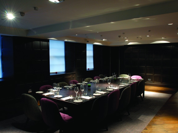 A view of one of the Blythwood's meeting spaces shows a dark panelled room with golden parquet flooring. Fabric blinds are pulled down over each of the 3 visible windows. The room is lit with spotlights in the white ceiling. A long dark-wooden table is surrounded by plush chairs in plum and taupe fabrics. The table is decorated with dinner place-settings.