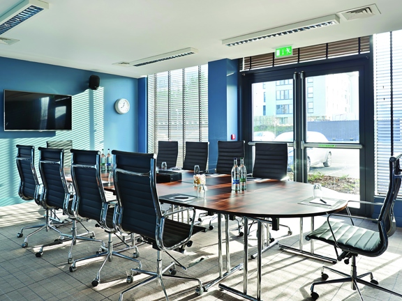 A photo of a board room style meeting room at Village hotel. The room has a beige, grid-patterned carpet and sky blue walls. The right hand wall is windowed, with a glass double door fire escape onto the carpark. The windows have pale venetian blinds down over them. There is a clock on the left wall beside a large wall-mounted TV screen and speakers can be seen. The large board room table has a wooden top and chrome legs and the 10 high-backed office chairs are in black leather with chromed bases. 