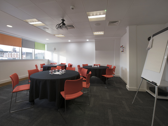 A meeting room at The Studio shows a corner of a room with white walls and dark grey carpet tiles. The left wall has 3 rectangle windows, side by side, with a view over the city; each one has a different coloured roller blind, in orange, grey and green. 3 round tables with black cloths are surrounded by red fabric chairs with wire legs. A projector is mounted on the tiled ceiling, between fluorescent lights. An A-frame flip chart is set up on the right-hand edge of the frame.