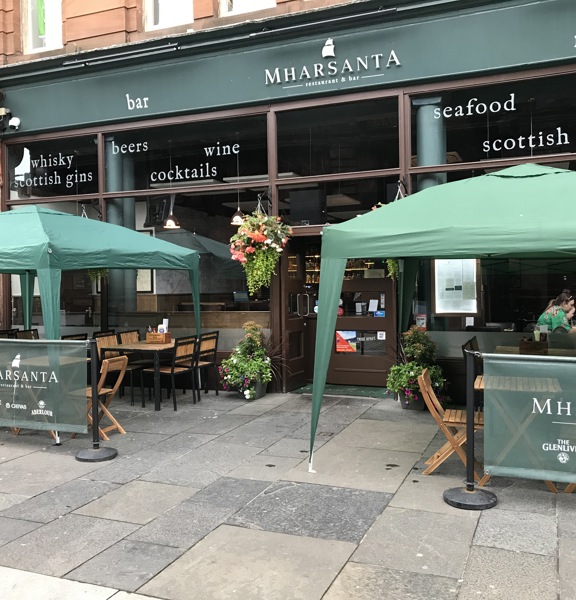 An exterior view of Mharsanta shows a restaurant with dark-green signage set into the ground floor of a red-sandstone building. The top panels of the windowed front is decorated with white wording describing the restaurants specialities, e.g. scottish classics, seafood and whisky. Outdoor, wooden seating is arranged behind dark green branded banners, under 2 gazebos, either side of the doorway. The pavement is wide, level and paved. A grand, carved stone doorway can be seen on the left-edge of the photo.
