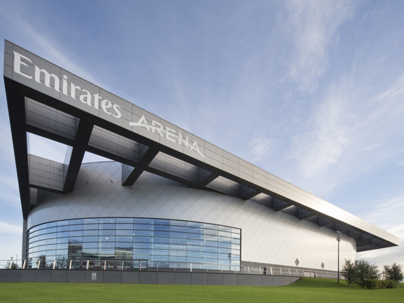 An exterior view of the Emirates Arena shows a modern rounded building with a large latticed structure adorning the roof. The closest curved wall is windowed, half the height of the building, while the rest is covered in silver tiling. The closest beam of the grey, latticed structure has large white lettering reading " Emirates Arena." The building is seen surrounded by a large lawn, small trees and a blue, cloudy sky.