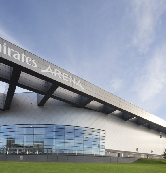 An exterior view of the Emirates Arena shows a modern rounded building with a large latticed structure adorning the roof. The closest curved wall is windowed, half the height of the building, while the rest is covered in silver tiling. The closest beam of the grey, latticed structure has large white lettering reading " Emirates Arena." The building is seen surrounded by a large lawn, small trees and a blue, cloudy sky.