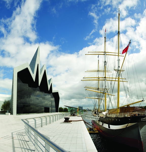 An exterior of the Riverside Museum. On the left, the museum is a modern silver building with a cartoonish, zig-zagging roofline. The main facade if a huge, dark glass wall which overlooks the Tall Ship and the River Clyde. Outside the building is a wide, pale stone, paved area and footpath with a silver railing between the building and the waters edge. A ramps leads to the Tall Ship is moored on the right, its hull is black and white and its multiple masts are bright yellow against the blue cloudy sky.