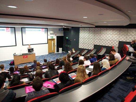 A university of Strathclyde lecture theatre, taken from the top & back of the tiered seating. The floor is carpeted tin a geometric pattern of shades of blue. A lecturer stands at a wooden desk at the front of the room with a monitor on top. 2 projectors beam images onto screens on he white wall behind them. Students sit dotted throughout the red, self-righting seats. A wooden internal door and a double doored, external fire escape can be seen in the far, front corner of the theatre.
