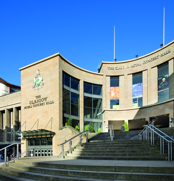 An exterior image of the venue. Curved steps with metal handrails lead up to the curved stone facade of the concert halls main entrance. The side of the building bears the Glasgow coat of arms and lettering reading, "The Glasgow Royal Concert Hall". The venue has 3 large doorways at the top of the stairs and floor to ceiling windows flank them over 3 storeys. There are 2 flag poles on top of the building seen against a clear, blue sky. Modern high-rise buildings can be seen to the left of the image.