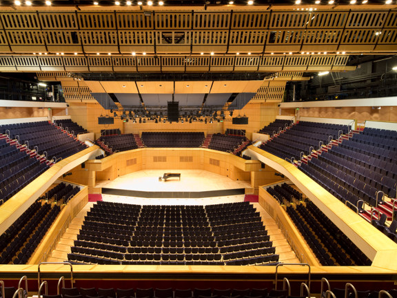 An interior image of GRCH's main auditorium from the back of the space, shows a stage surrounded by tiered seating over 3 levels. The stage and its surrounds are light wood while the carpeting and seating are furnished in deep red and black respectively. Lights can be seen across the ceiling and throughout the auditorium. The ceiling appears almost metallic and grilled, technical lighting and sound equipment are evident.