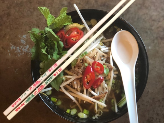 A close up, top-down view of a bowl of Hanoi Bike Shop's pho soup. A black bowl is filled with broth,  noodles, beansprouts, spring onions, red chilli, mint and other garnishes. A white soup spoon and chopsticks are perched on the bowls rim.